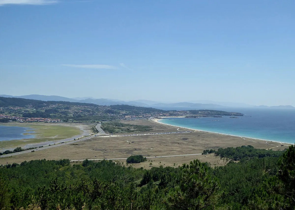 Vista elevada del istmo de A Lanzada: una franja plana de dunas y pradera con aguas interiores a la izquierda y la playa con el mar turquesa a la derecha