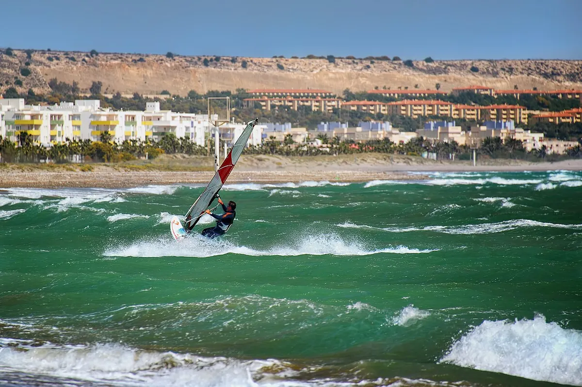 Windsurfista sobre olas de agua verde frente a Almerimar, con edificios blancos y un cantil al fondo