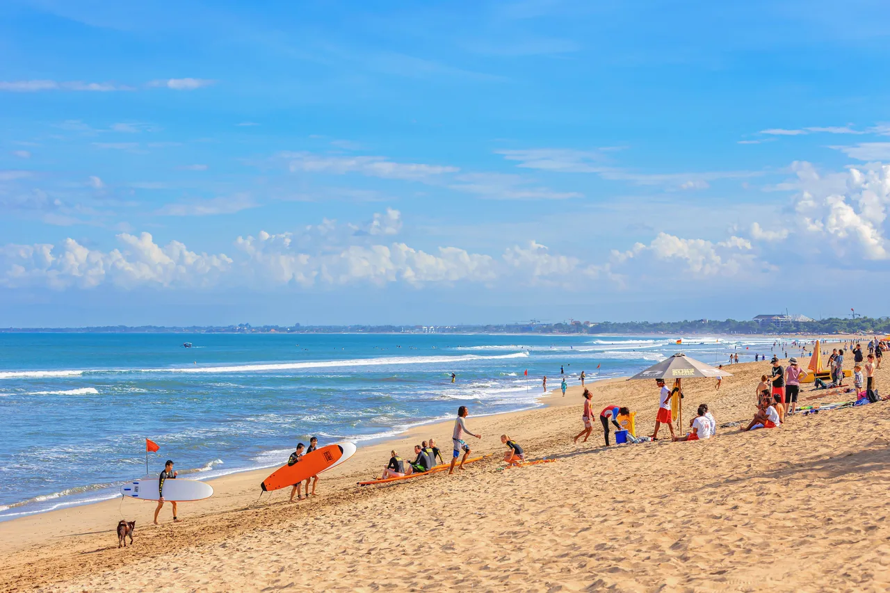 Surfers carrying soft-top boards and a group sitting by their boards on a long sandy beach break in south Bali, with a small red flag at the water's edge
