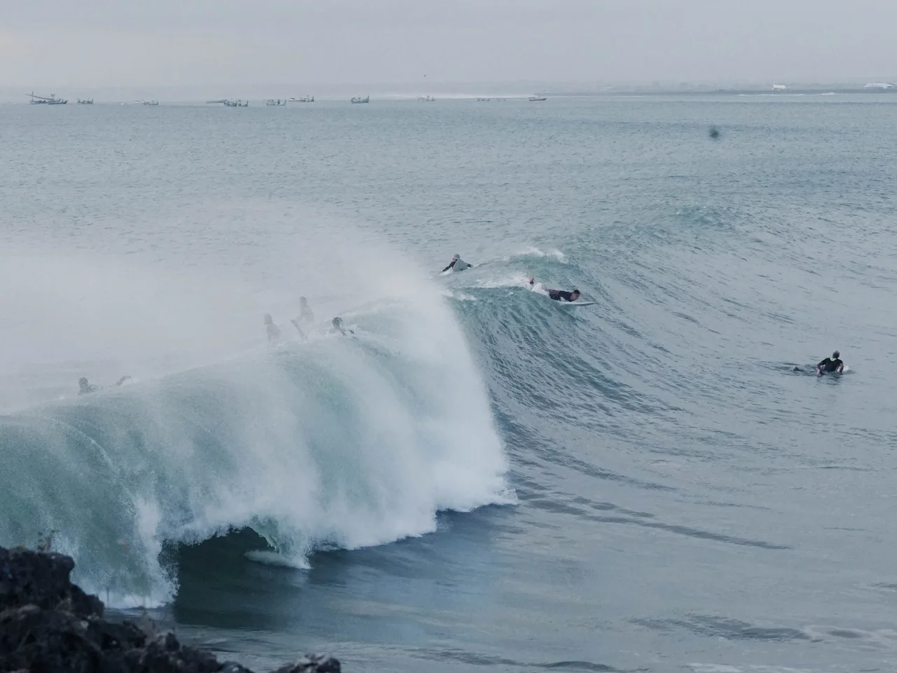 Surfers paddling over and riding a heavy reef wave below dark rocks in South Kuta district, Bali