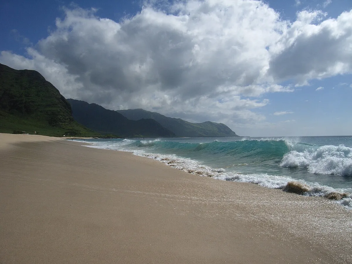 Vague qui déferle sur une plage de sable, avec des montagnes verdoyantes à l'arrière-plan