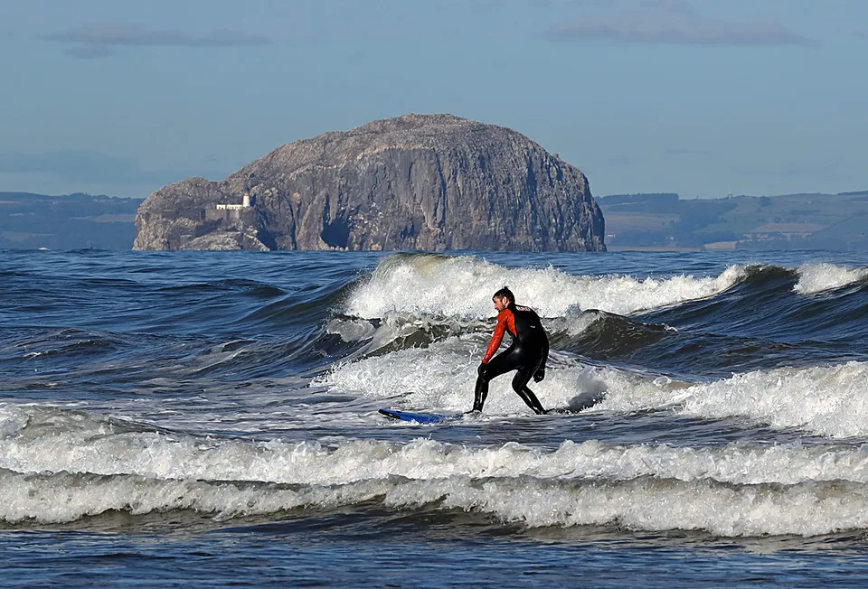 A surfer riding white water at Belhaven Bay with the Bass Rock behind