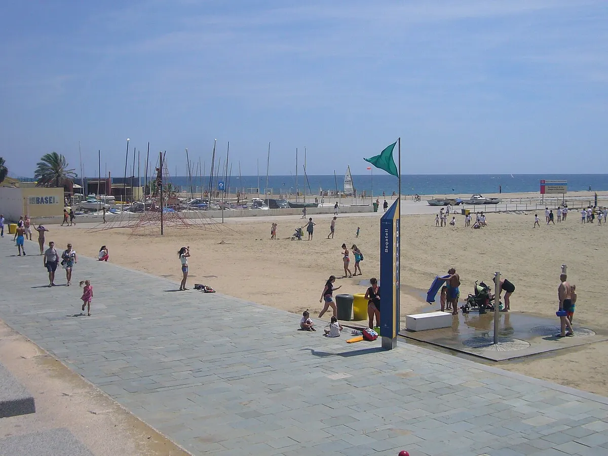 Promenade and sand at Bogatell beach in Barcelona, with bathers and boat masts in the background