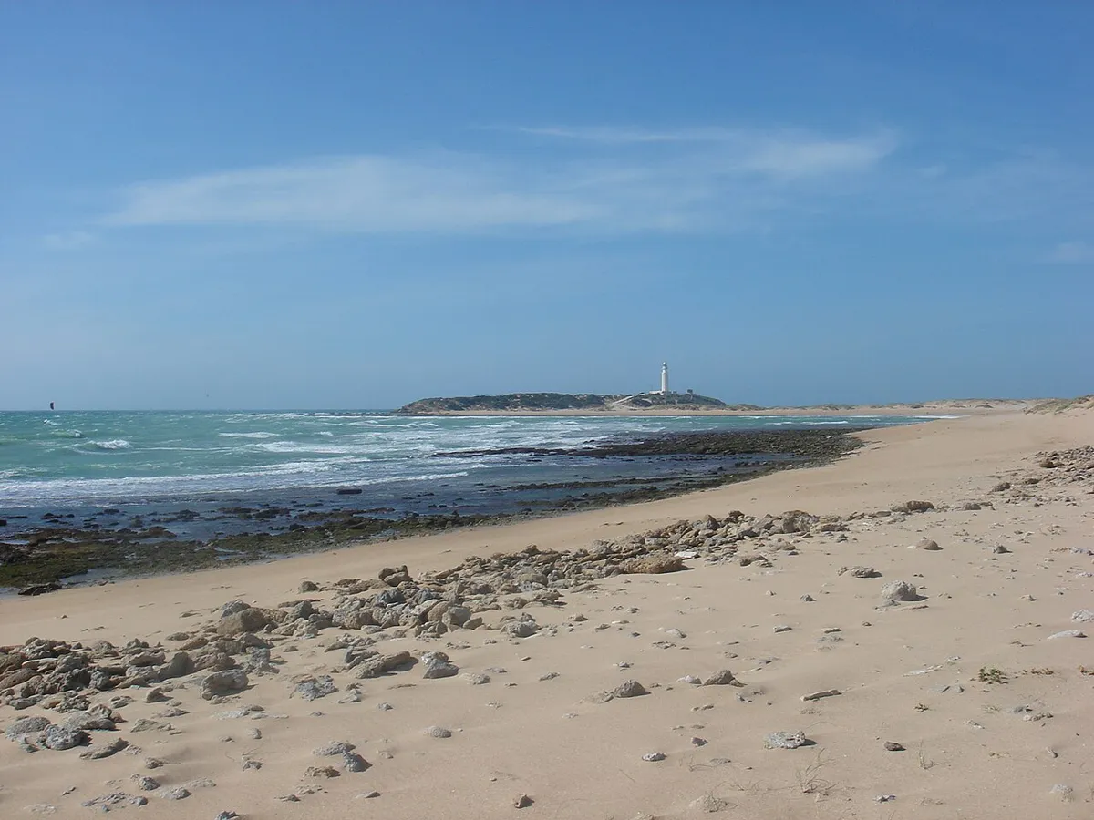 Orilla de arena y rocas en Los Caños de Meca, con el faro de Trafalgar en el horizonte
