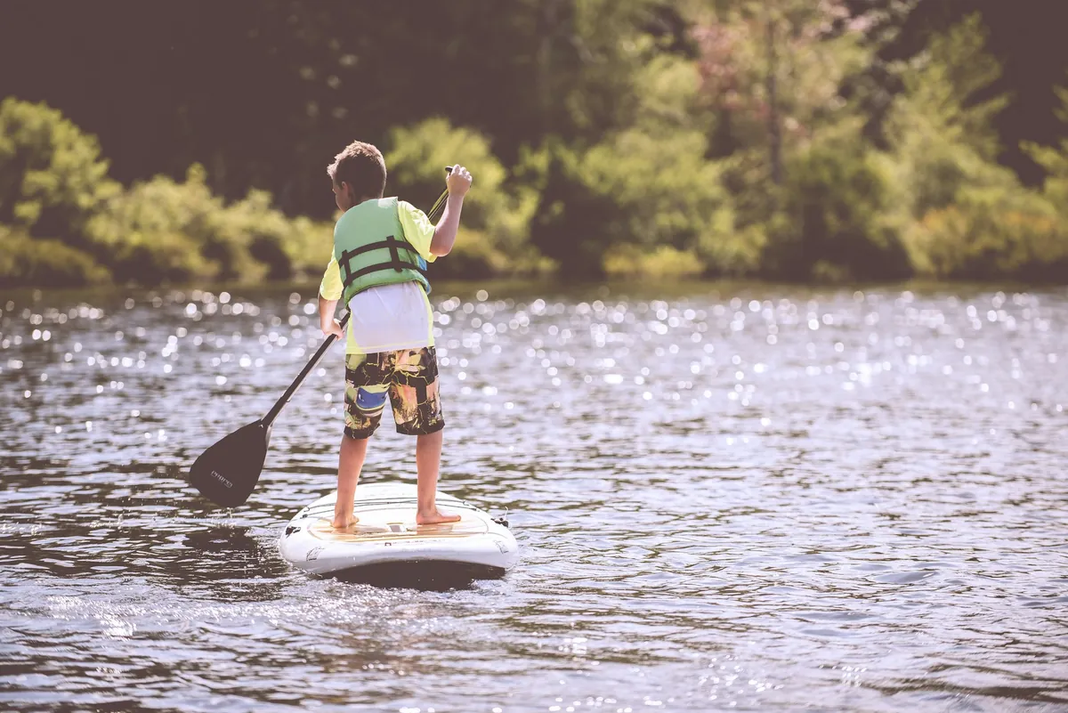 Un enfant debout sur une planche de SUP, pagaie en main, portant un gilet de flottabilité jaune et vert