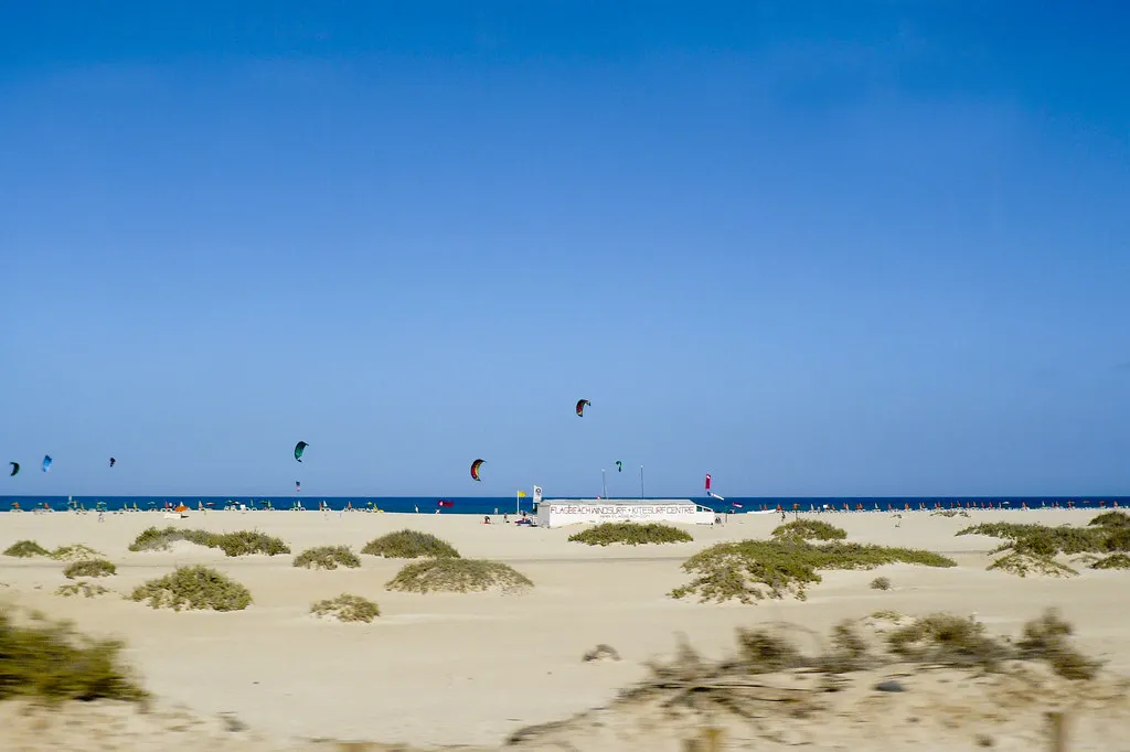 Kites in the air above a sandy beach with a low building signed Flagbeach Windsurf + Kitesurf Centre, at Flag Beach, Corralejo