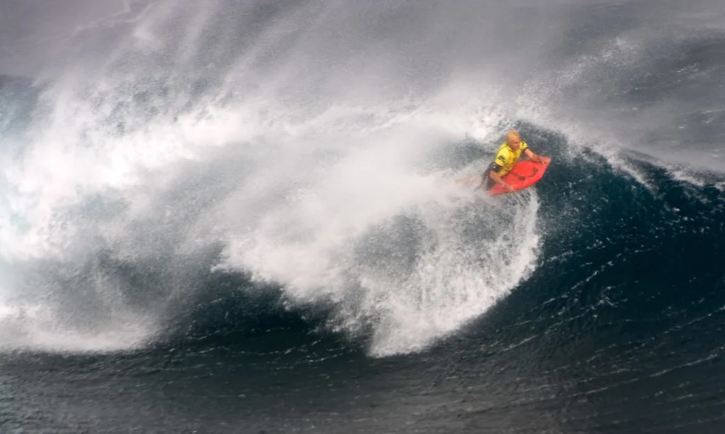 Bodyboarder in a yellow top on a red board riding the face of a large wave, with spray thrown up behind