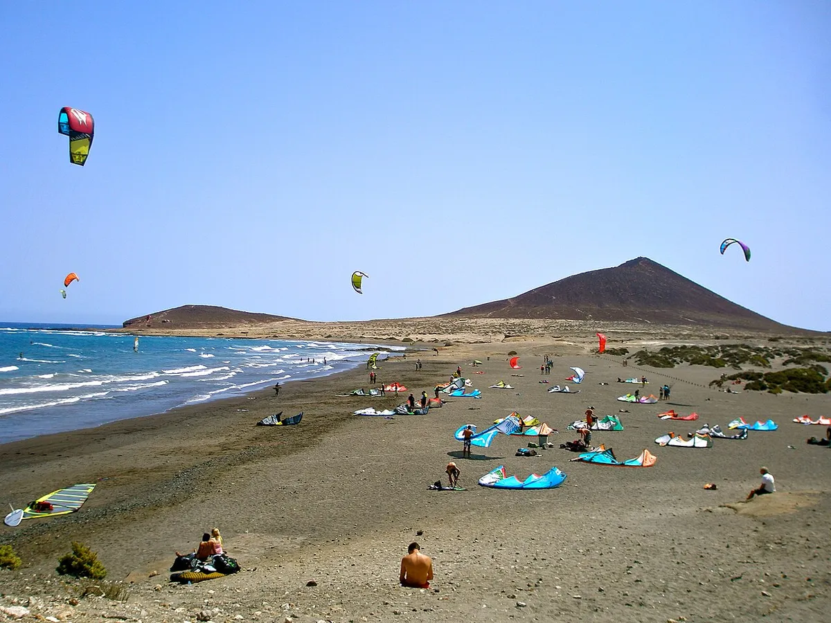 Kites in the air and kite gear laid out on a dark-sand beach at El Médano, with a conical hill behind