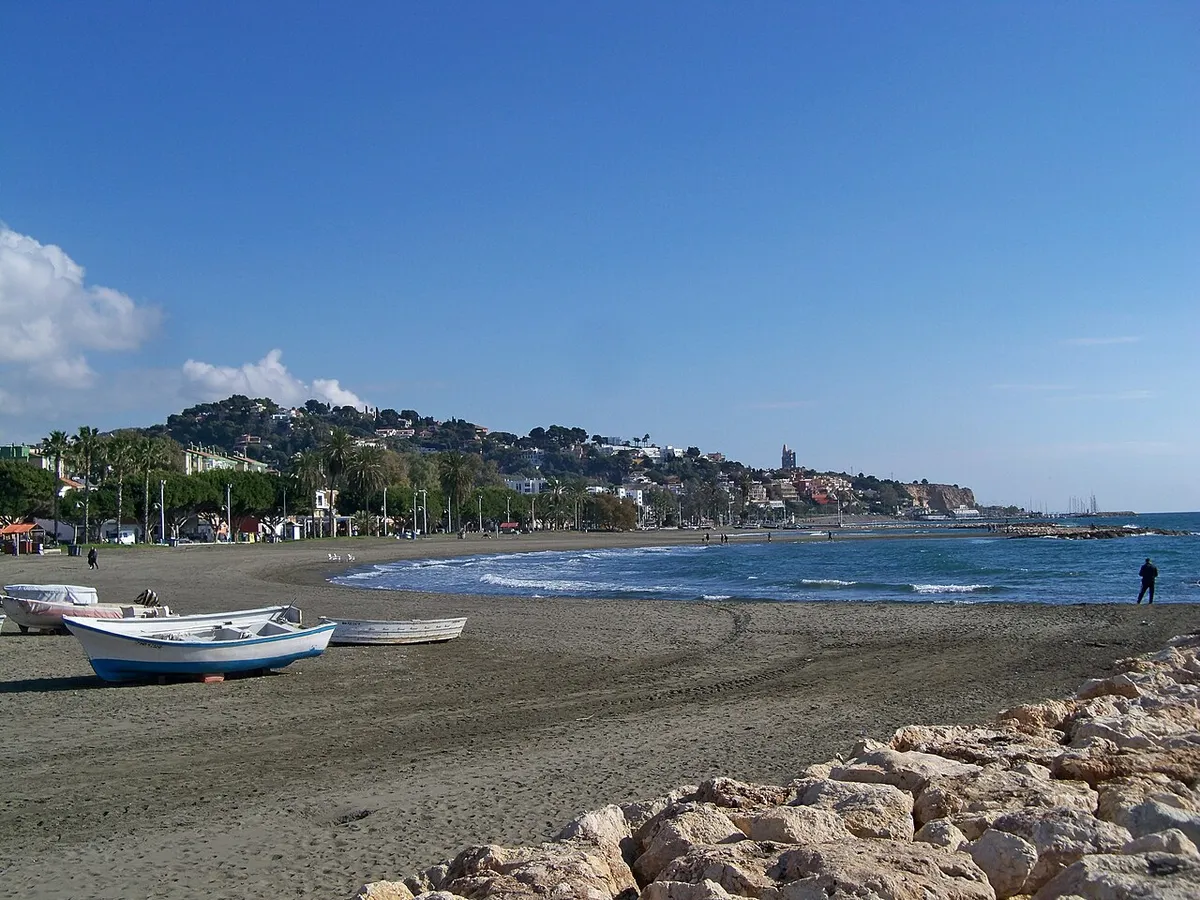 Playa de arena oscura de El Palo, en Málaga, con barcas varadas en la orilla y las colinas de la costa al fondo