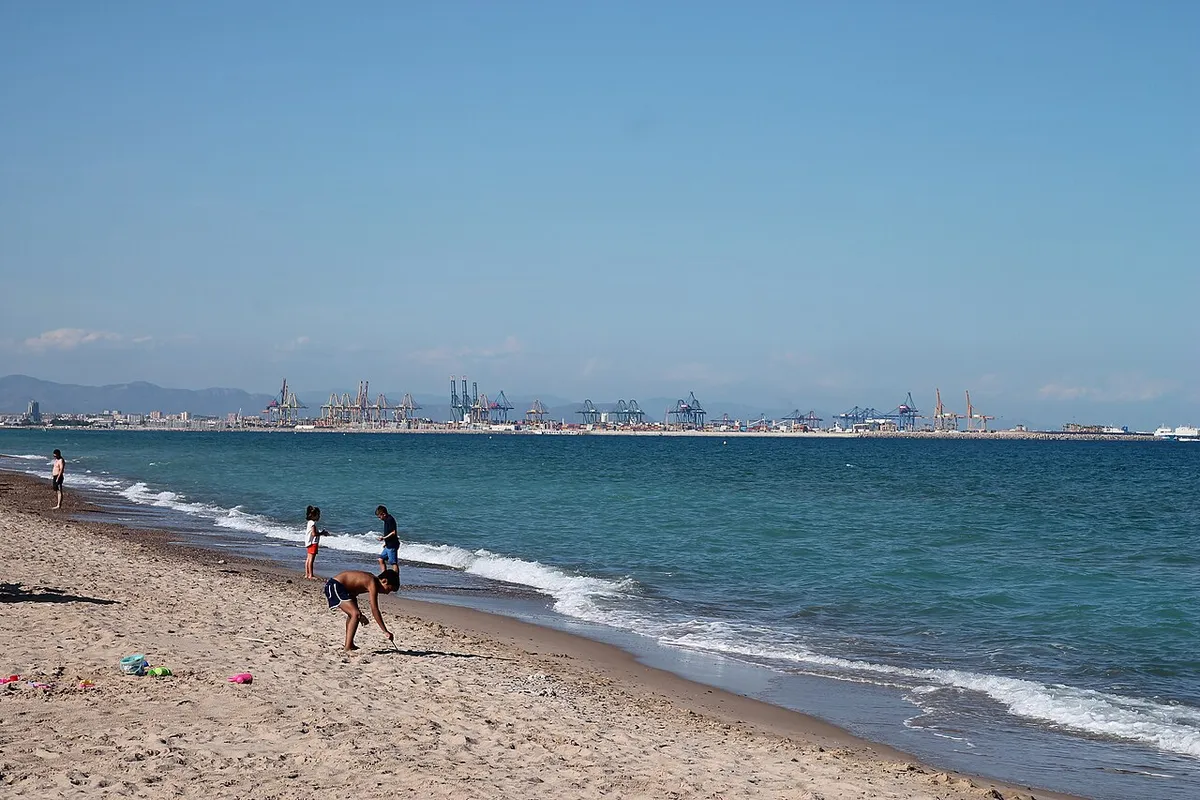 Orilla de arena con oleaje y bañistas en la playa de El Saler, en Valencia, con las grúas del puerto al fondo