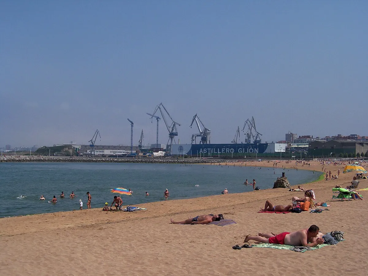 Playa de El Arbeyal, en Gijón, con bañistas en la arena, aguas tranquilas y grúas al fondo