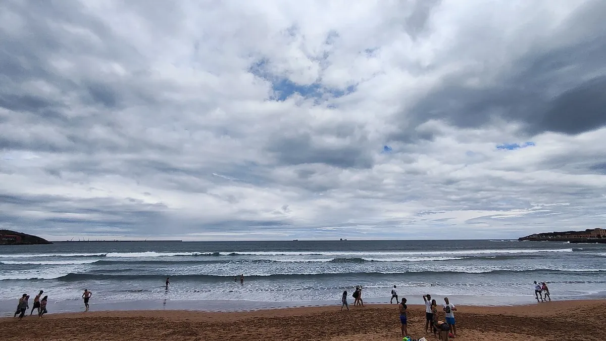 Olas rompiendo en la orilla de la playa de San Lorenzo, en Gijón, con gente sobre la arena bajo un cielo nublado