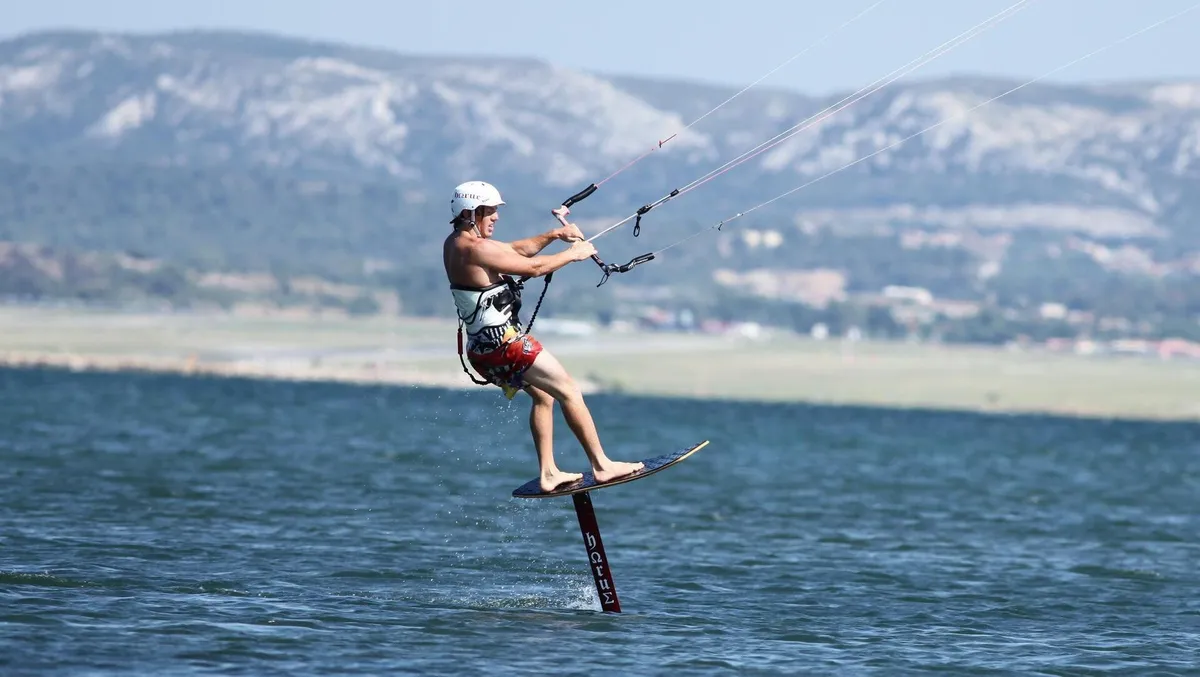 Pratiquant de kite foil sur sa planche, soulevée au-dessus de l'eau par un mât, avec des montagnes en arrière-plan