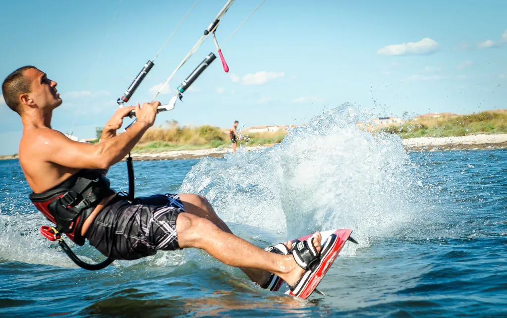 Kitesurfeur penché en arrière sur la carre de sa planche, dans une gerbe d'eau, la barre en mains