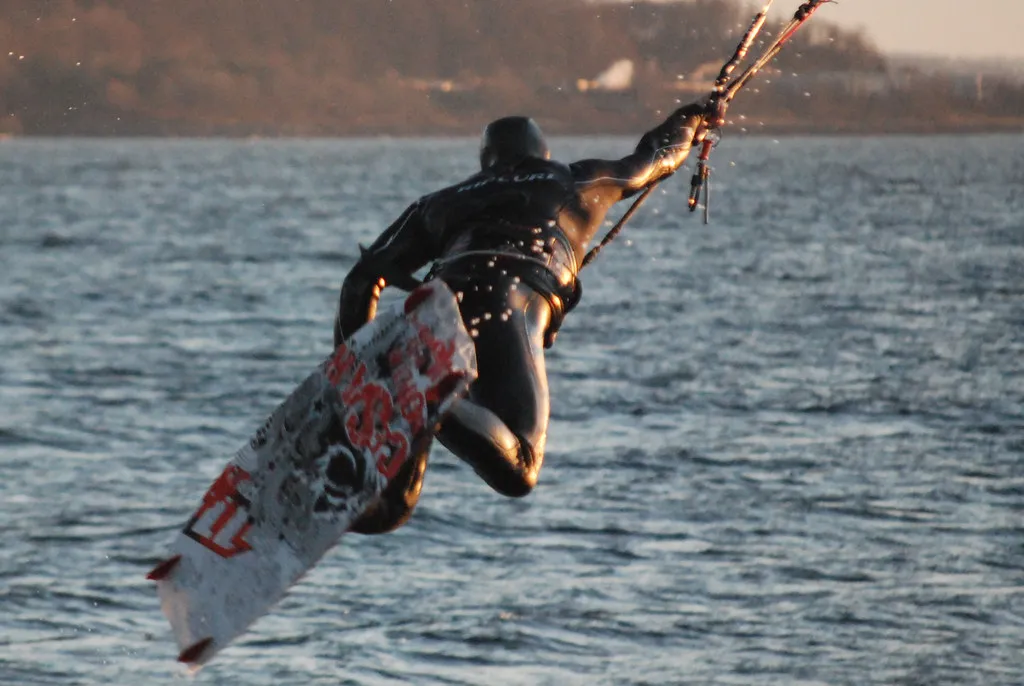 Kitesurfer in a black full-length hooded wetsuit, mid-jump, with one hand on the bar and the board below
