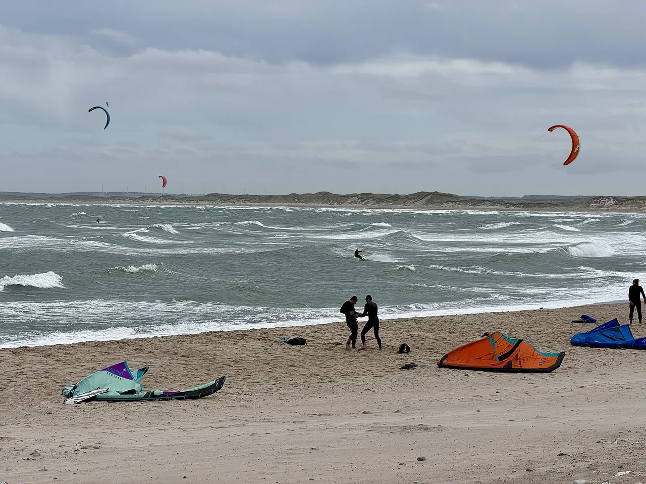 Kitesurfers rigging on the sand at Klitmøller beach while others ride the waves under grey skies