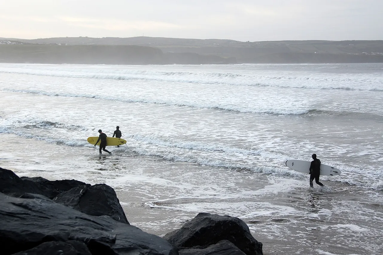 Three surfers carrying longboards out of the shallows at Lahinch, with rocks in the foreground and the far side of the bay behind