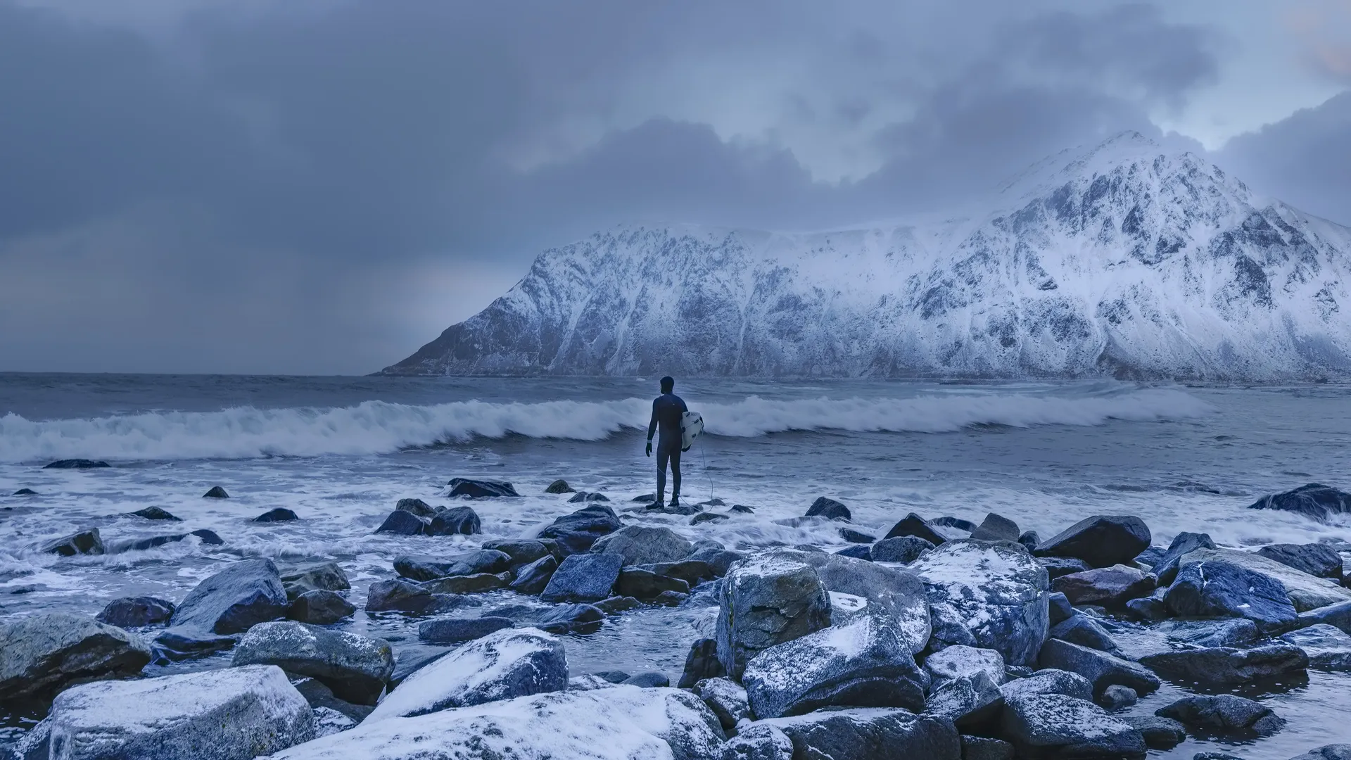 A surfer in a wetsuit standing on snow-covered rocks in the Lofoten Islands, facing breaking waves and a snowy mountain