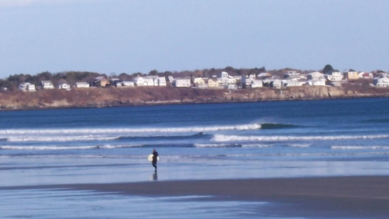 A surfer carrying a board along Long Sands Beach, York, Maine, with Cape Neddick and small waves in the background