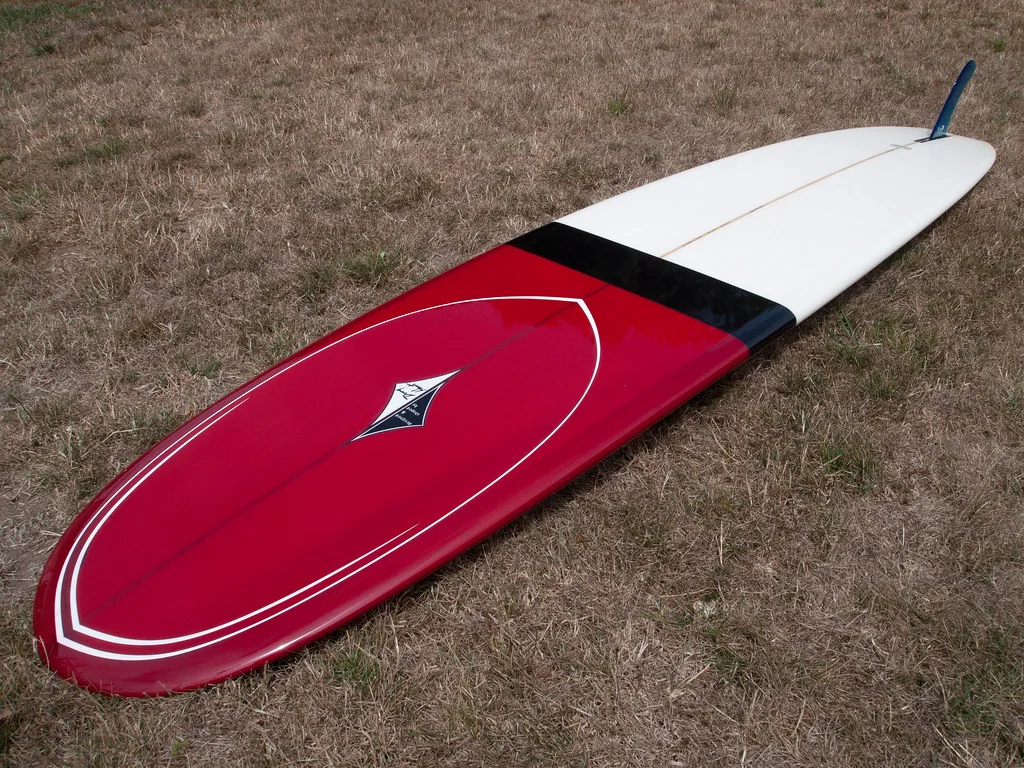 Longboard rouge et blanc à nez arrondi et à dérive unique, posé sur l'herbe
