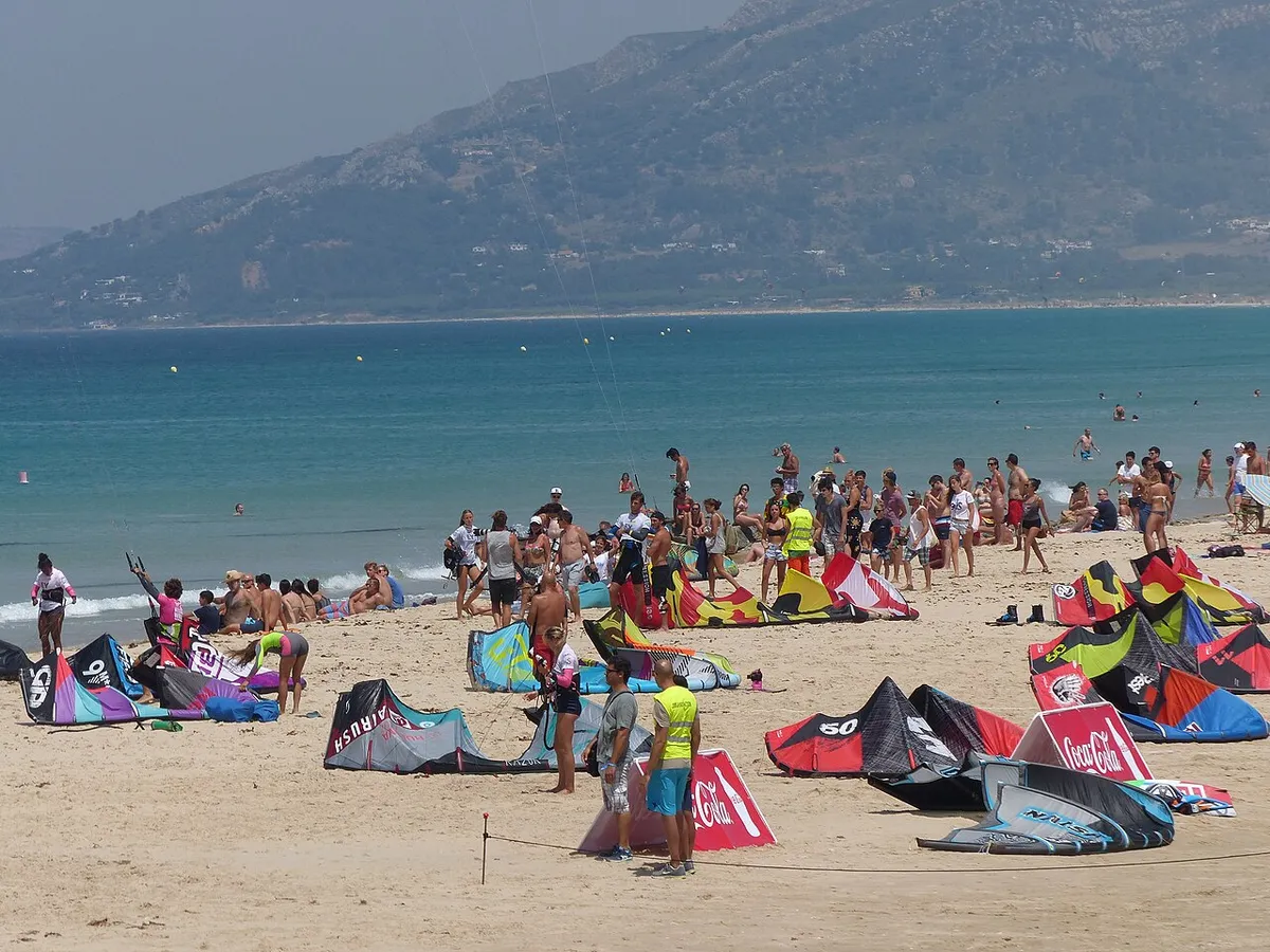 Kites déployés sur le sable de la plage de Los Lances à Tarifa, avec des kitesurfeurs et des montagnes en arrière-plan