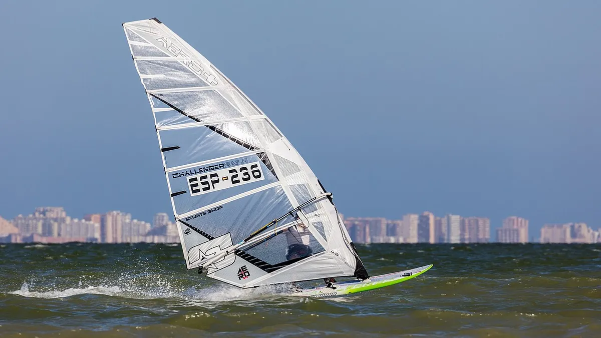 Windsurfista con la vela blanca inclinada sobre el agua del Mar Menor, con una hilera de edificios altos en el horizonte