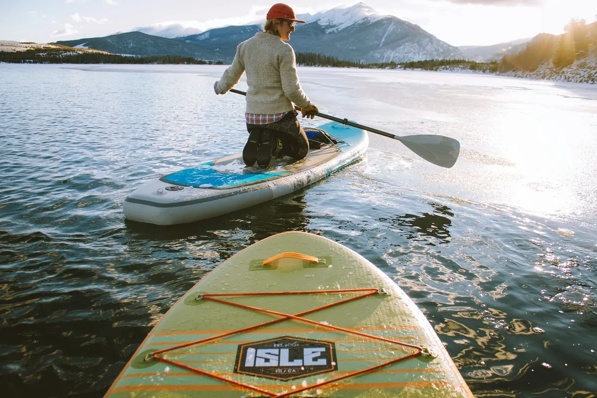 Une personne à genoux sur un paddle sur un lac de montagne au soleil bas, avec l'avant d'une seconde planche au premier plan