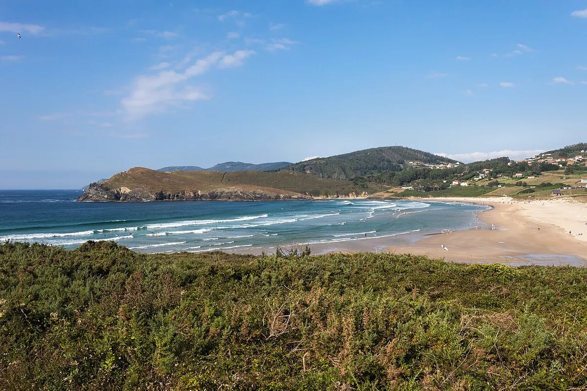 Vista de la playa de Pantín, en Valdoviño (Galicia): líneas de olas rompiendo hacia una playa de arena, con un promontorio verde a la izquierda y casas a la derecha