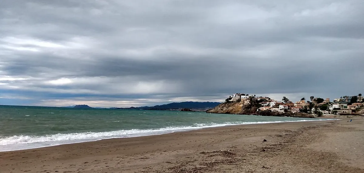 Playa de arena con oleaje bajo un cielo cubierto en Bolnuevo, Mazarrón, con casas sobre un promontorio al fondo