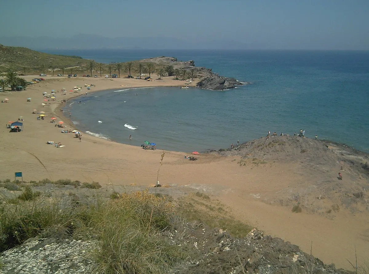 Pequeña cala de arena entre cerros bajos con el mar azul, en la playa de Percheles