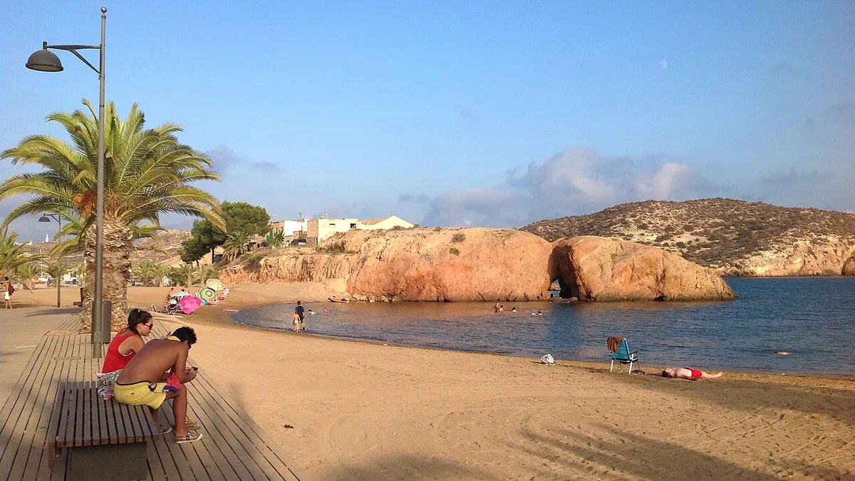 Paseo de madera y palmeras junto a la playa de arena de la Ermita, con grandes rocas rojizas en el agua