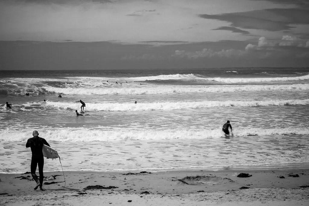 Photo en noir et blanc de surfeurs dans les vagues à la Pointe de la Torche, avec un surfeur en combinaison qui marche vers l'eau, planche sous le bras
