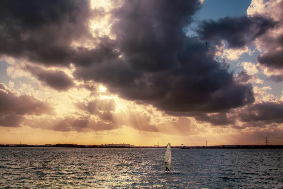 A windsurfer on the water under a sunset sky with rays of light breaking through the clouds