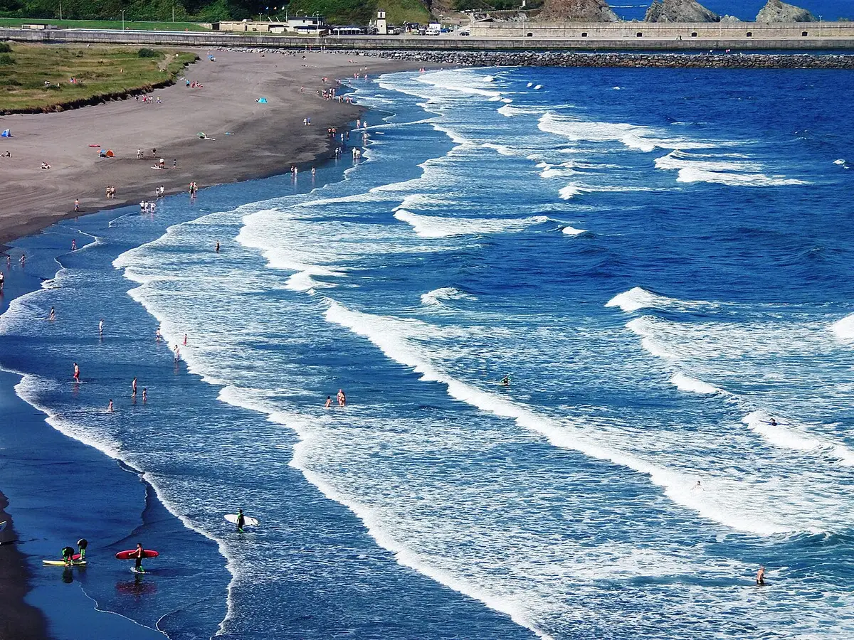 Plage de La Arena (Asturies) vue d'en haut : des courants d'arrachement apparaissent comme des trouées dans l'écume et des bandes d'eau plus sombre entre les vagues qui déferlent