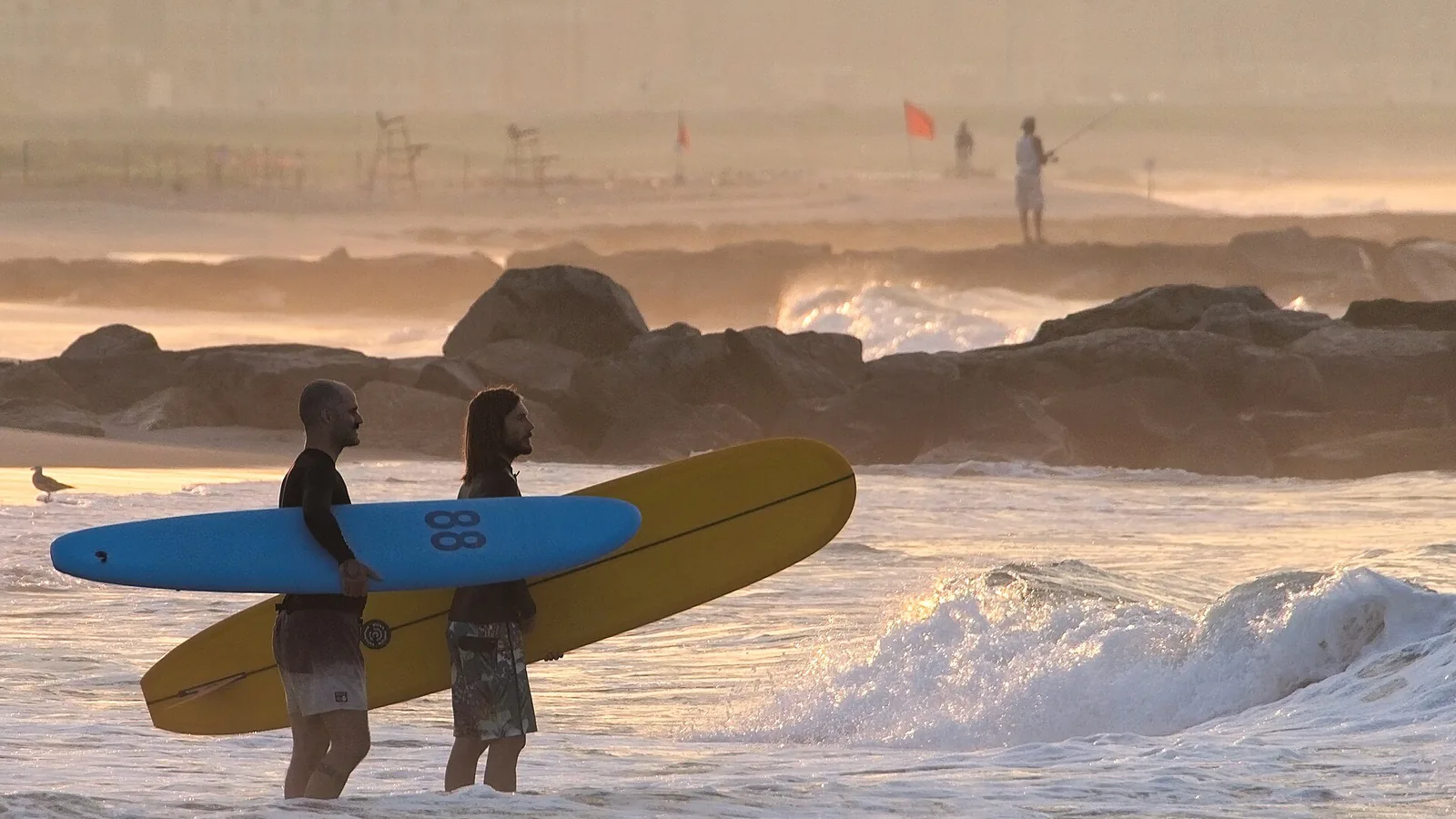 Two longboarders wading out at dawn beside a rock groyne at Rockaway Beach, New York