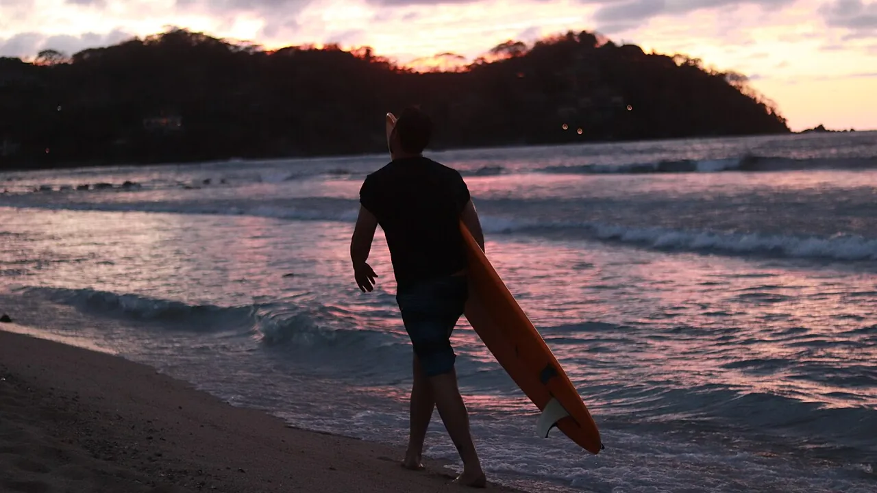 A surfer carrying a board along the shore at dusk in Sayulita, Nayarit, Mexico