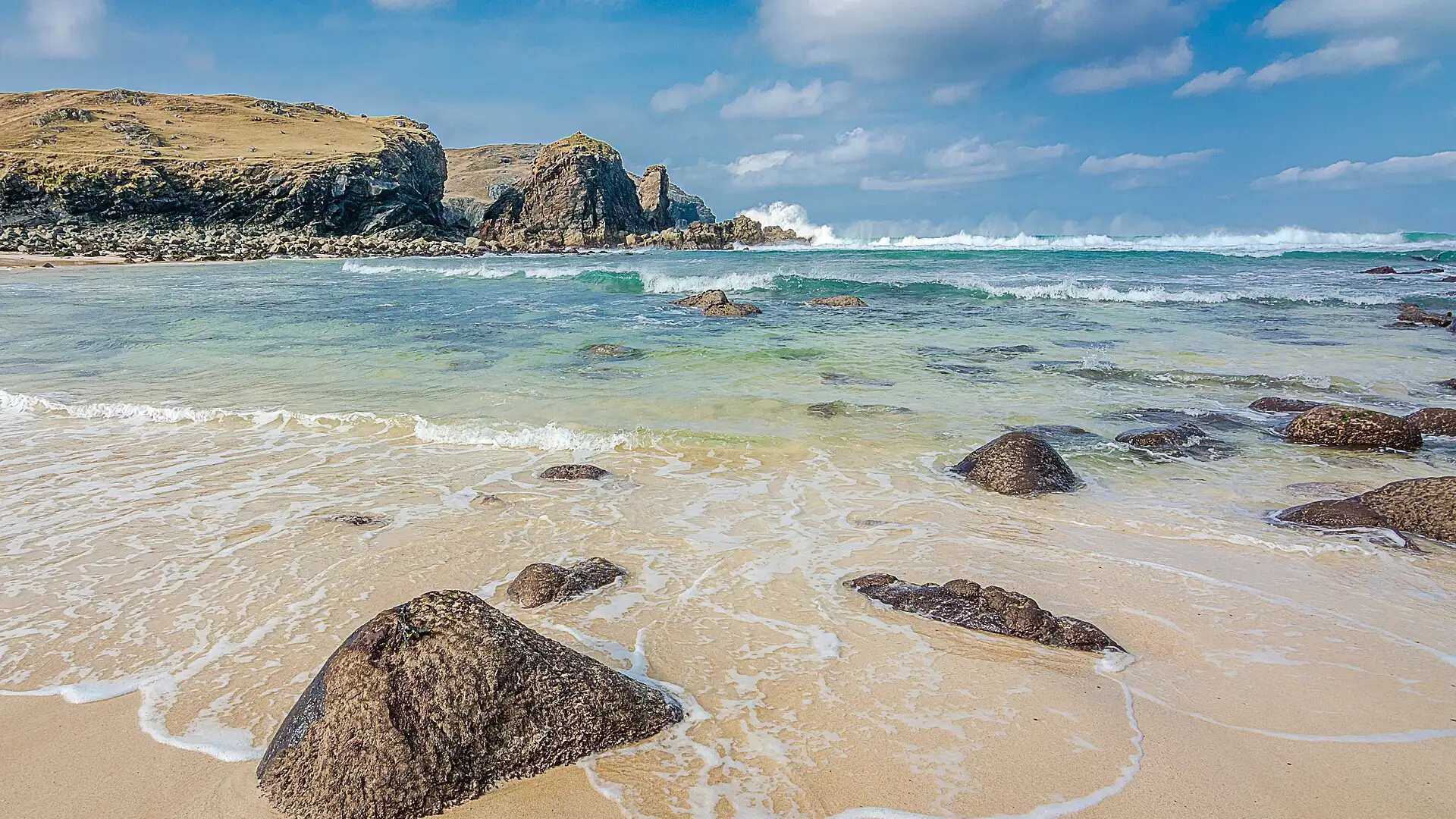 Waves breaking beyond a sandy beach scattered with boulders at Dalbeg, on the Isle of Lewis, under a blue sky