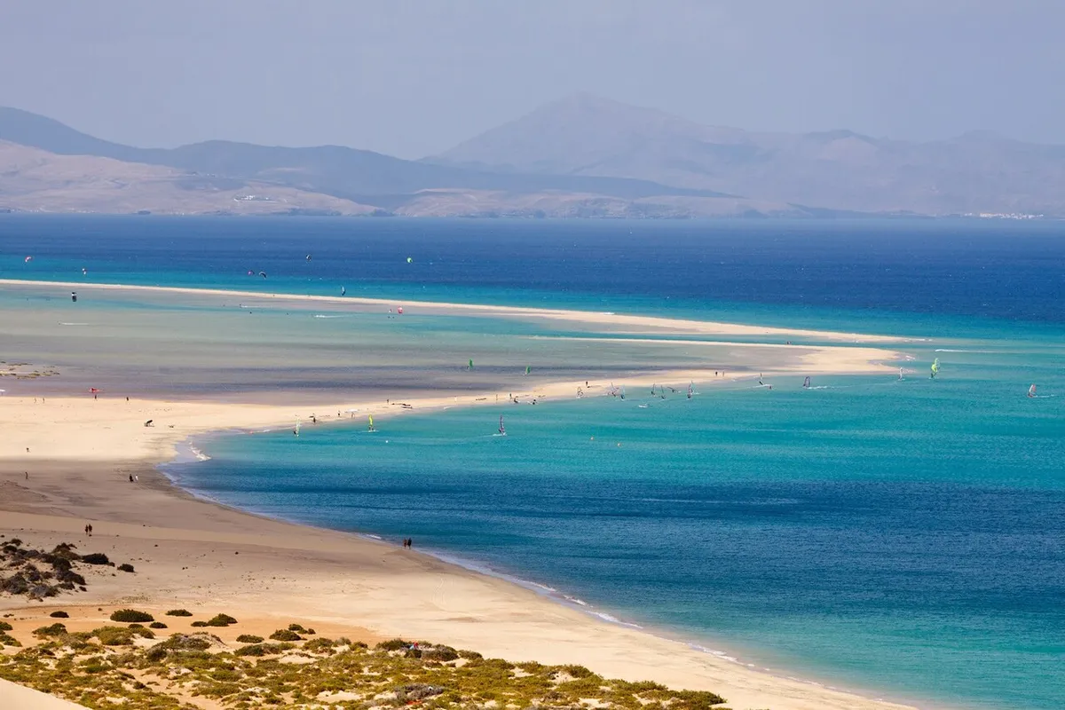 Vue en hauteur de la plage de Sotavento, à Fuerteventura : langue de sable et lagon turquoise parsemé de voiles de windsurf et de kitesurf