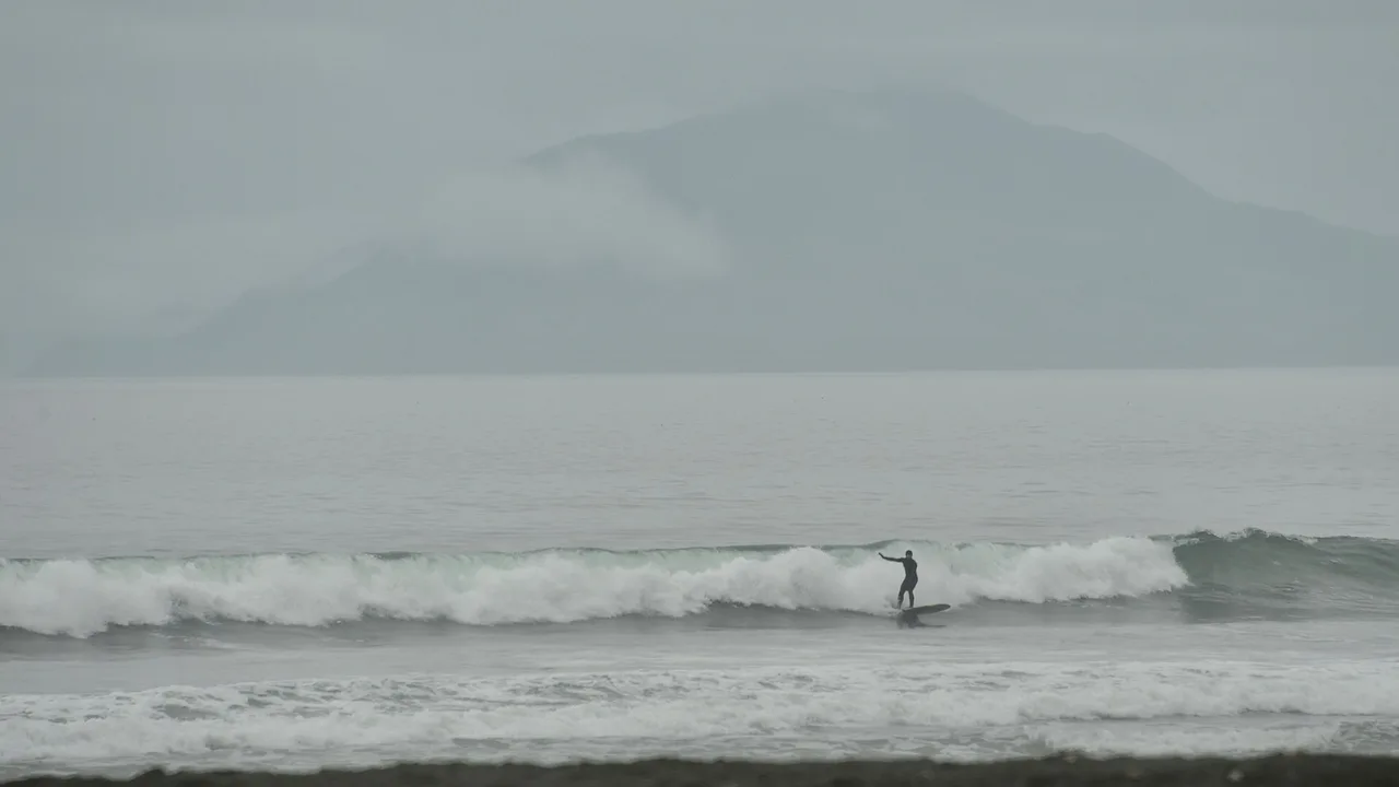 A surfer riding a wave at Surfers' Beach, Kodiak Island, Alaska, with a mountain in low cloud across the bay