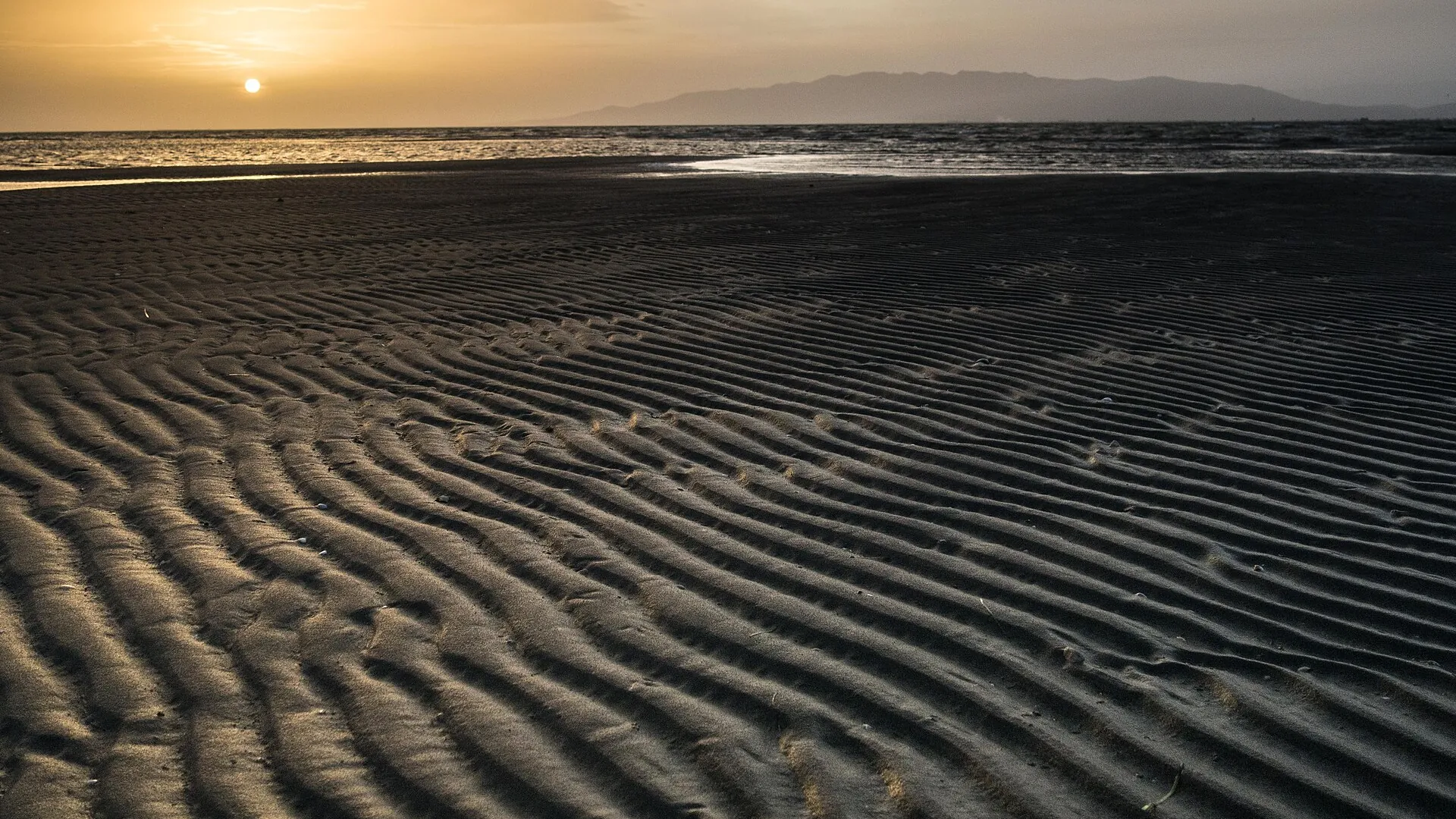 Rippled sand and the sea on the Barra del Trabucador in the Ebro Delta, with the low sun and distant hills