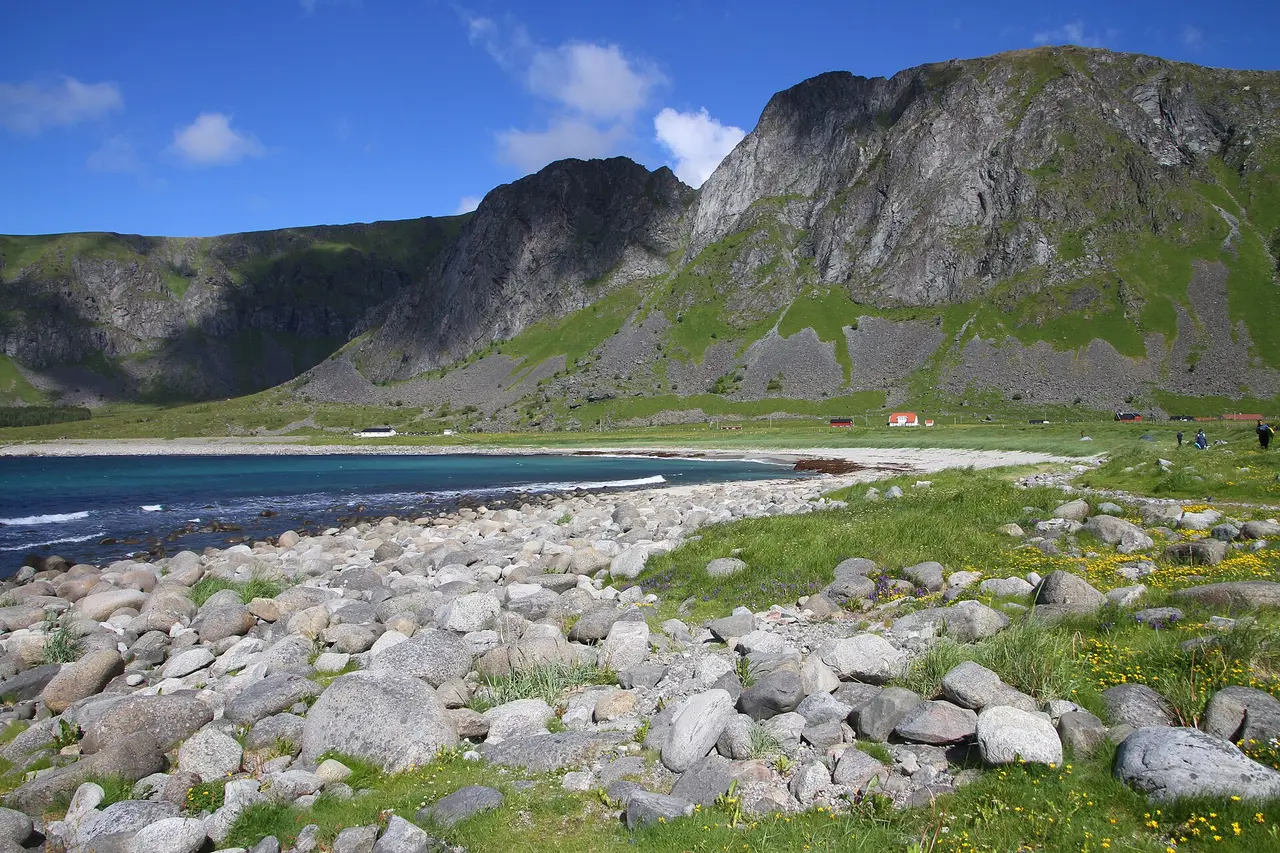 Unstad bay in summer: a stony shore in the foreground, a sandy beach and a few red buildings at the foot of steep green and grey mountains