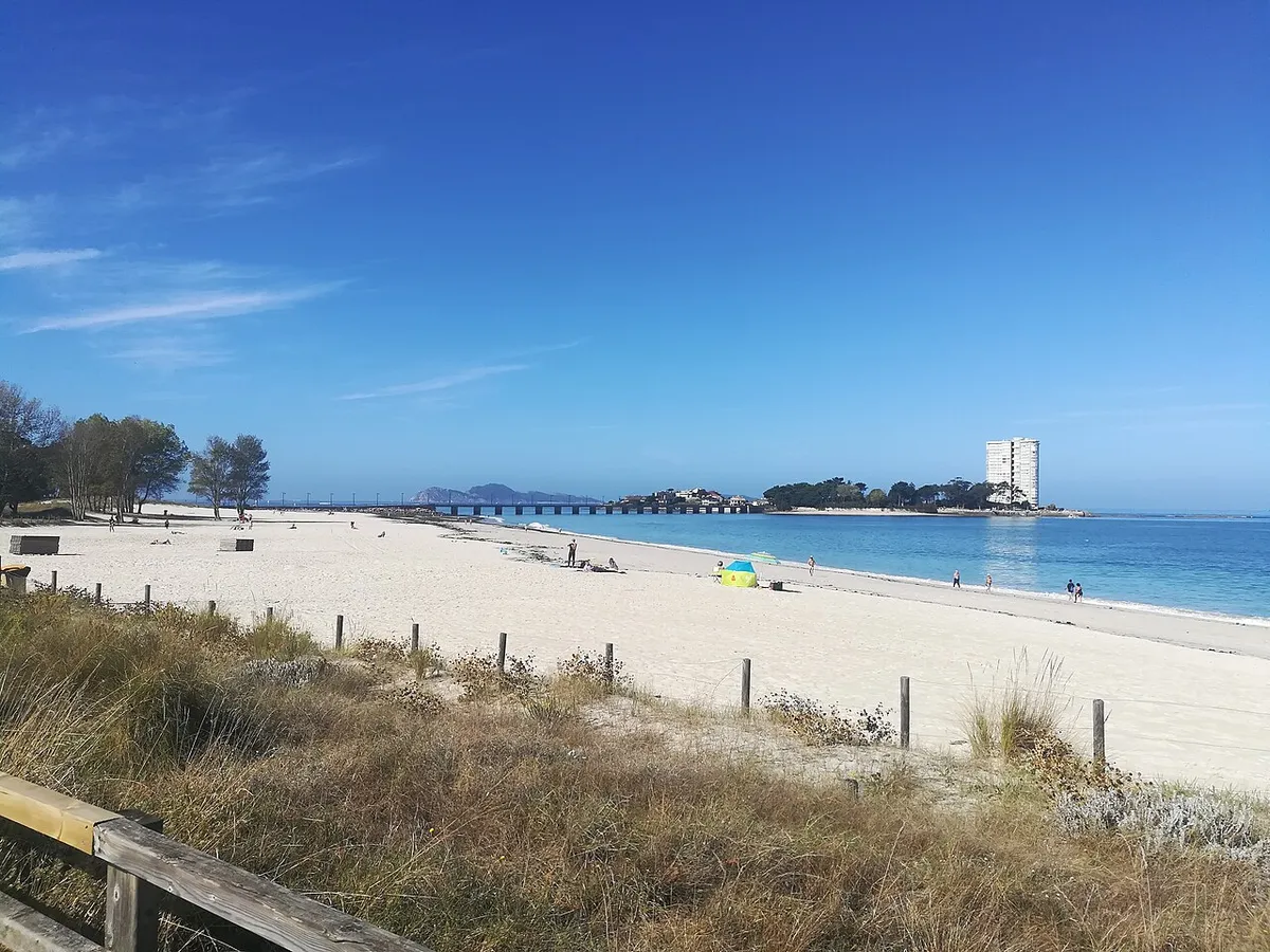 Praia do Vao, en Vigo: arena clara, dunas con vallas de madera y, al fondo, el mar con una isla y un edificio alto