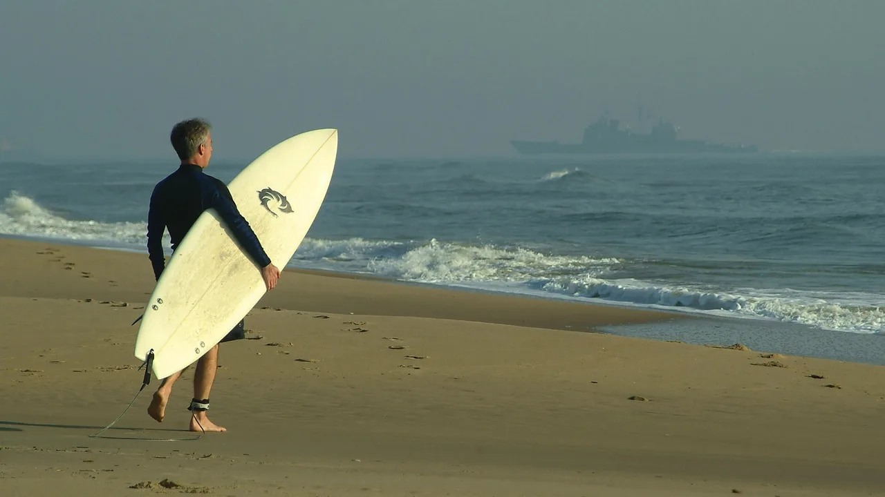 A surfer carrying a board along the beach at Virginia Beach, Virginia, with a Navy warship visible offshore