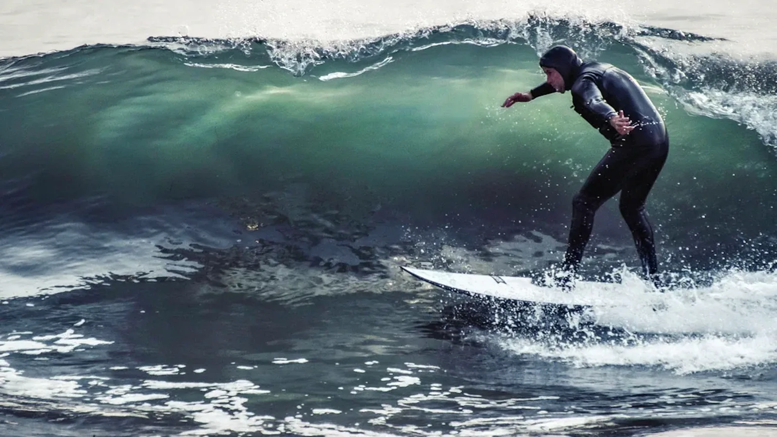 A surfer in a hooded winter wetsuit riding a cold-water wave