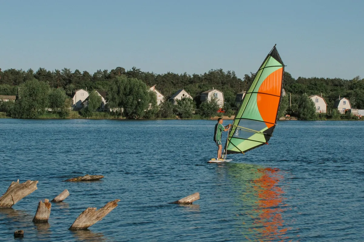 A windsurfer standing on the board holding the sail on a calm lake lined with trees and houses