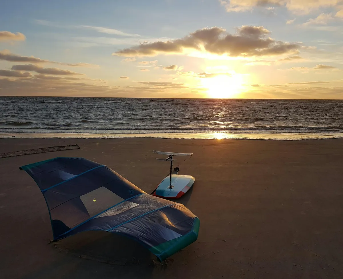 Ala de wing foil sobre la arena y tabla de foil de pie detrás, al atardecer