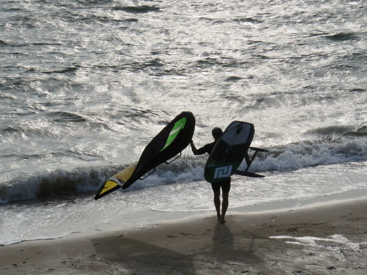 Rider de dos portant une aile et une planche à foil vers le bord de l'eau