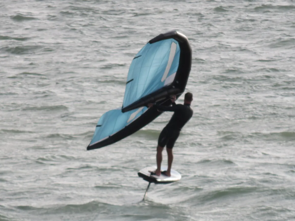 Rider debout sur sa planche à foil, tenant une aile bleu clair haut à côté de lui