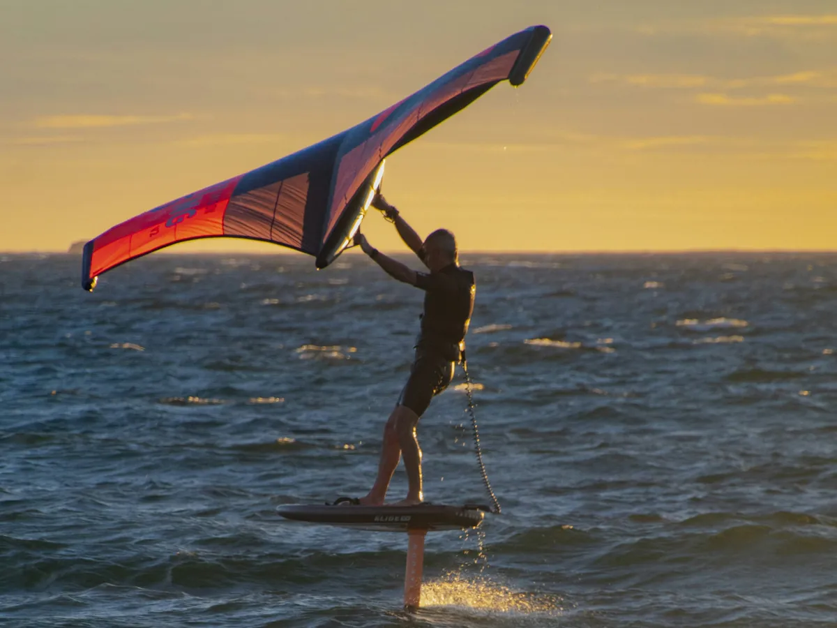 Rider de wing foil sur son foil, aile tenue haut au-dessus de lui, au coucher du soleil
