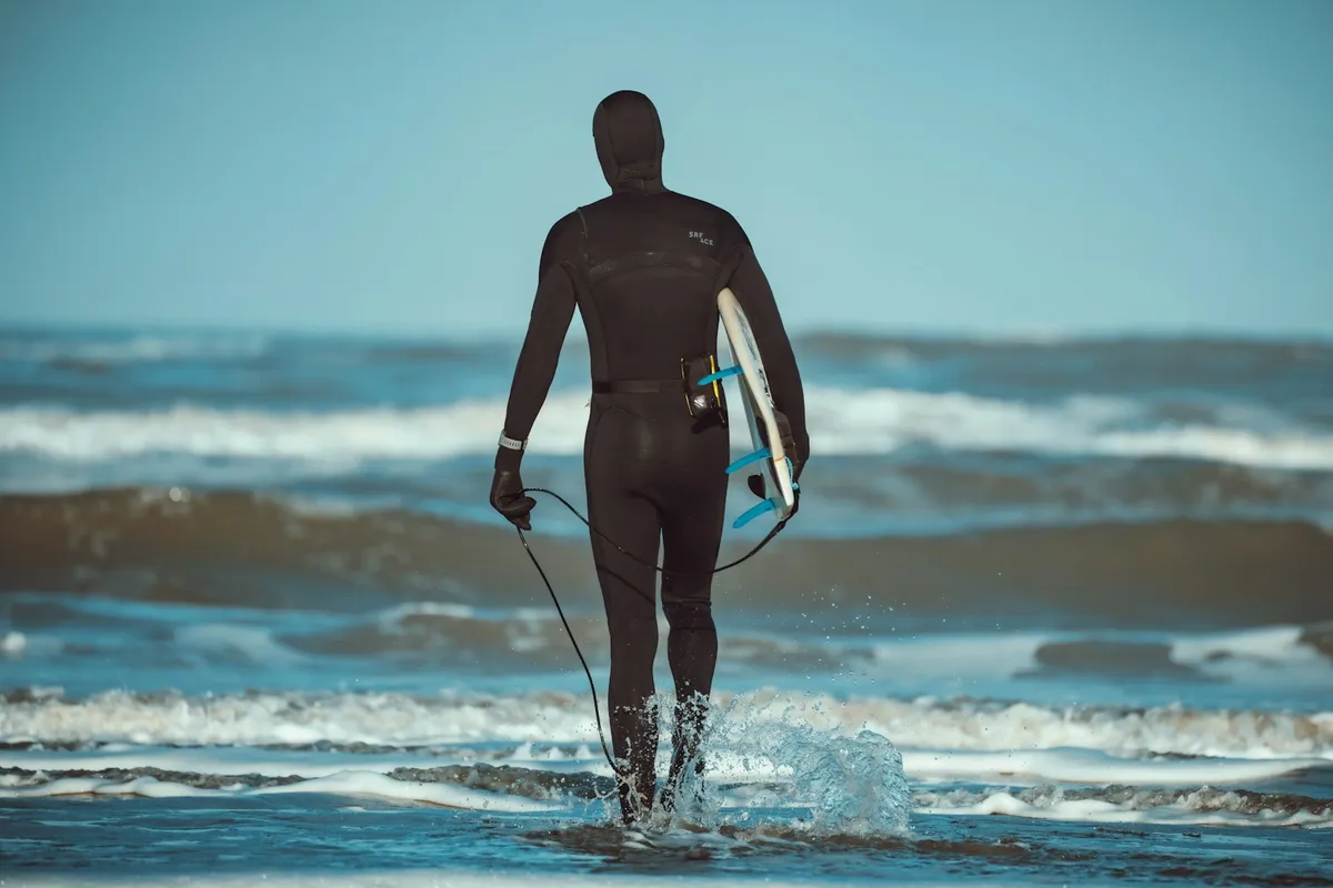 Surfeur vu de dos entrant dans la mer en combinaison intégrale noire avec cagoule et gants, planche sous le bras